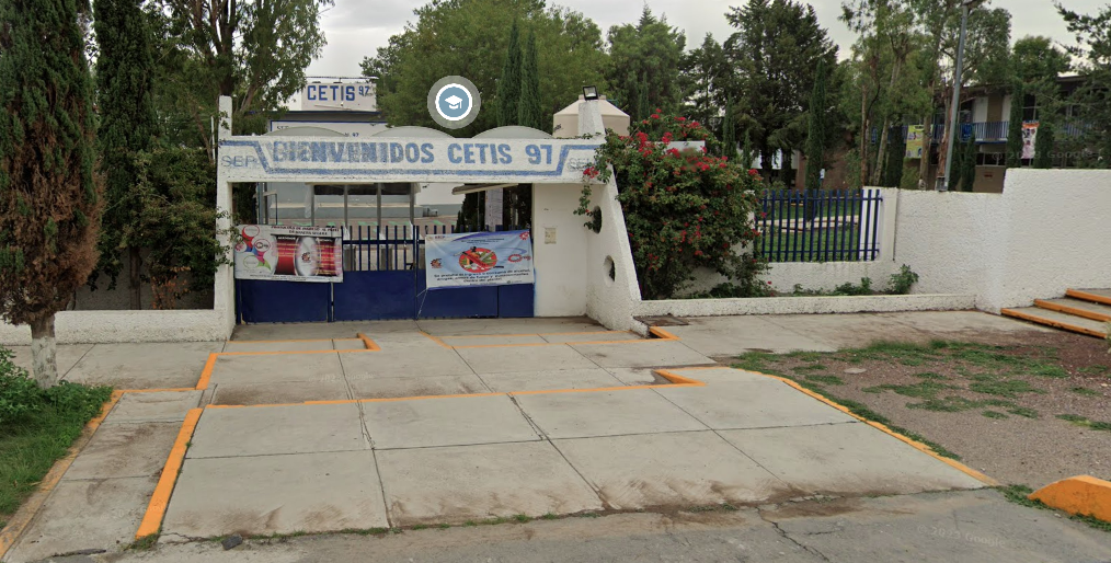 Fotografía en la que se muestra una explanada y al fondo hay una reja de color azul. Sobre la reja hay un muro blanco que tiene pintadas letras azules que dicen BIENVENIDOS CETIS 97.